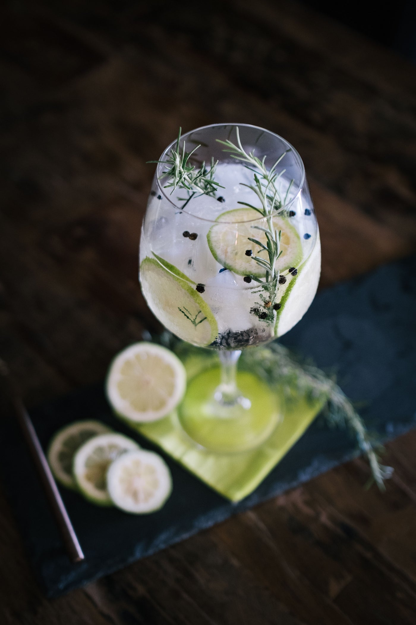 Gin and tonic garnished with lime, rosemary and juniper berries on a dark timber bench