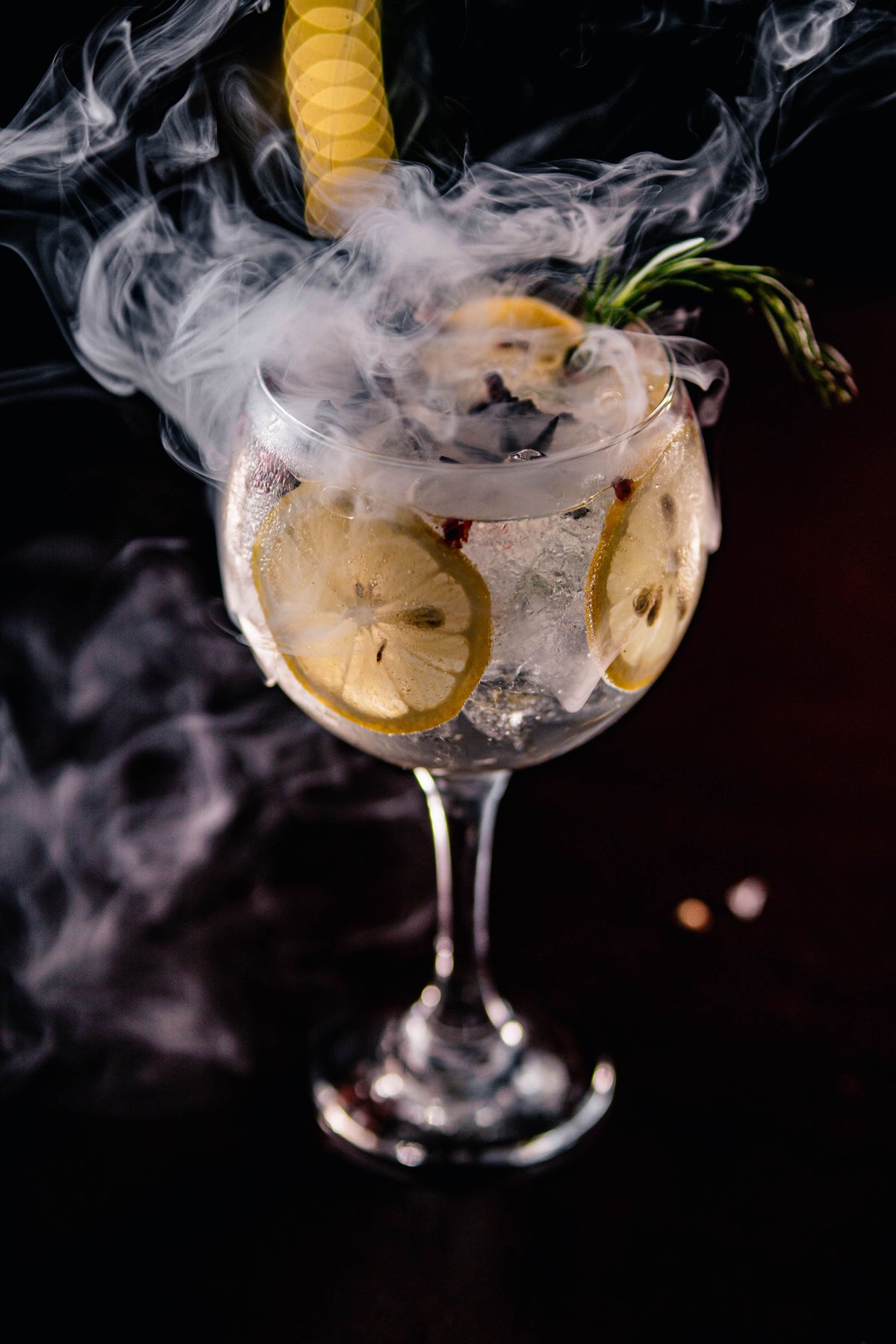 Gin and tonic in a copa glass with lemon and rosemary, wreathed in smoke against a dark background